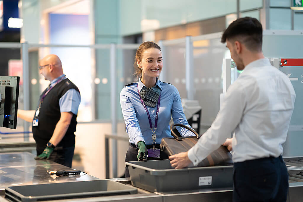 T5 Security performing different tasks. Security staff help passengers with checks on personal belongings in trays at a security area in Terminal 5 at London Heathrow Airport.