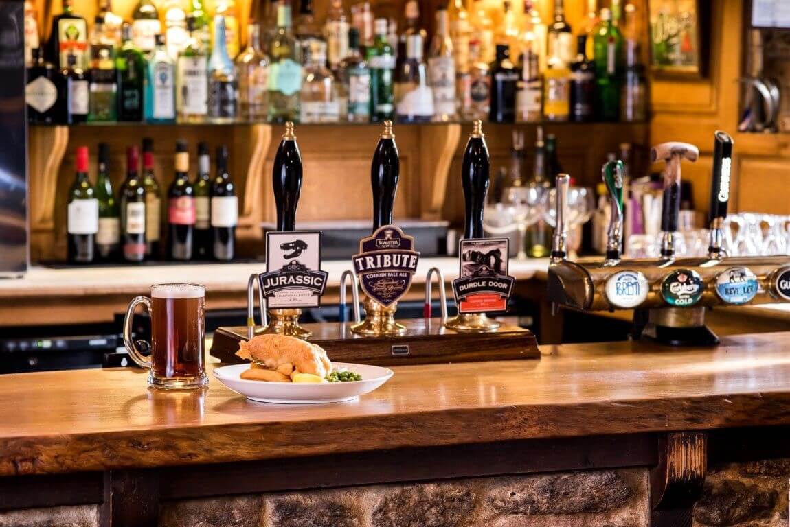 The bar displaying the pump handles for the draft ales and the spirits and wines in soft focus behind, with a plate of fish and chips and a pint of beer in a jug at The Acorn Inn, Dorset. VisitEngland Awards for Excellence 2024 Finalist