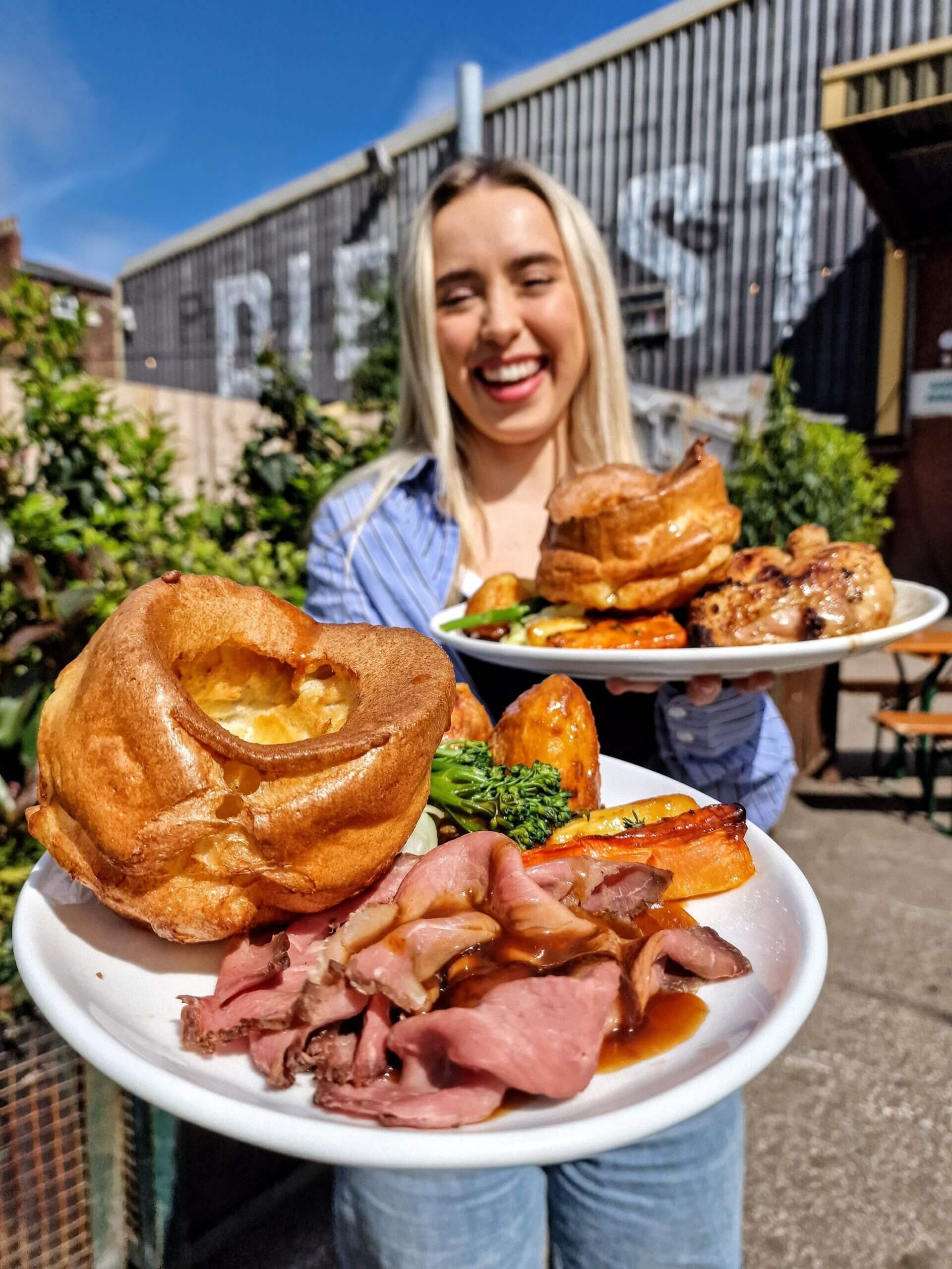 A smiling woman holding large plates of pub food in Diecast - a large creative space, music venue and beer garden in Manchester.