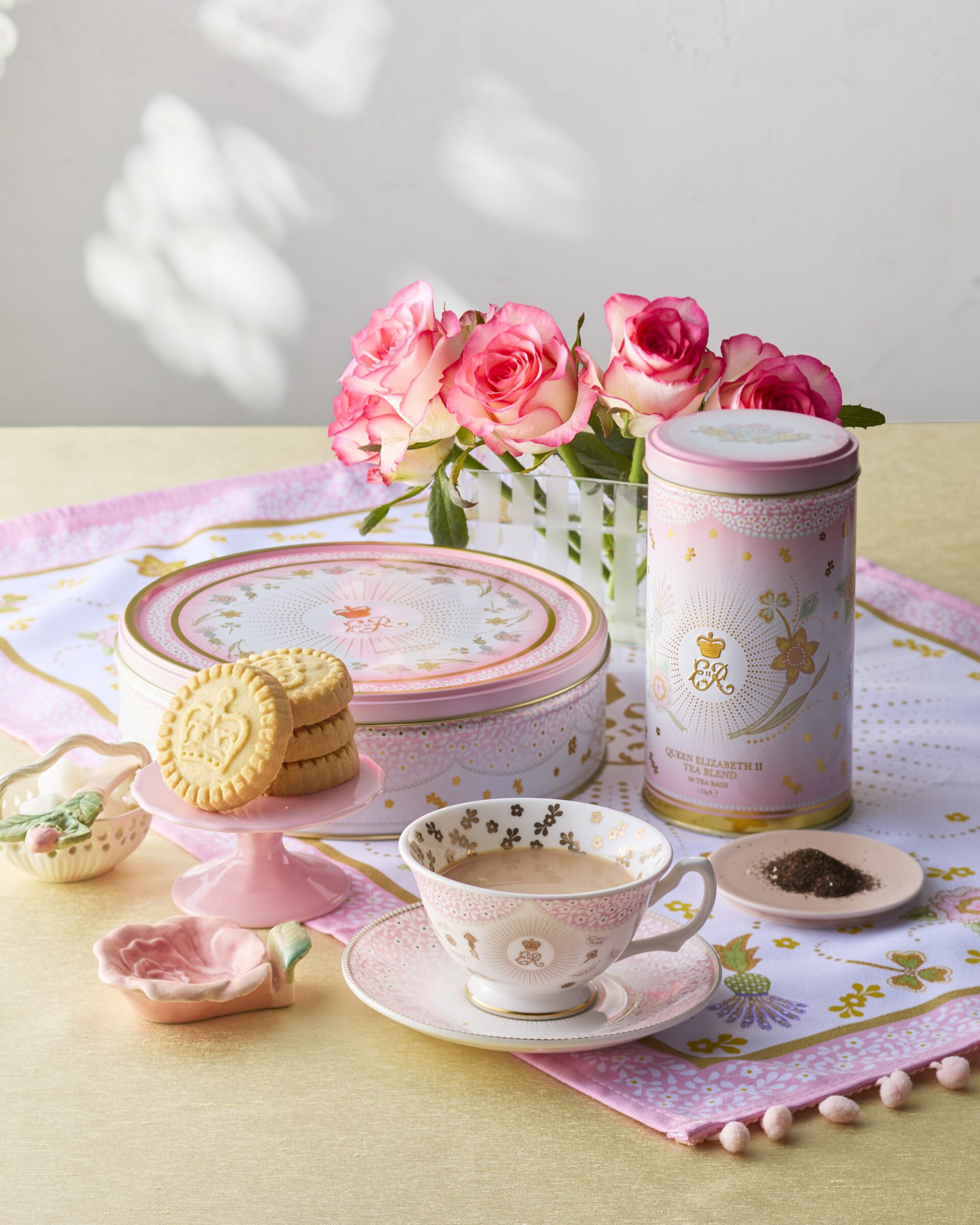 バラの花とともに並ぶエリザベス2世女王陛下100周年記念の紅茶缶やビスケット缶とティーカップ