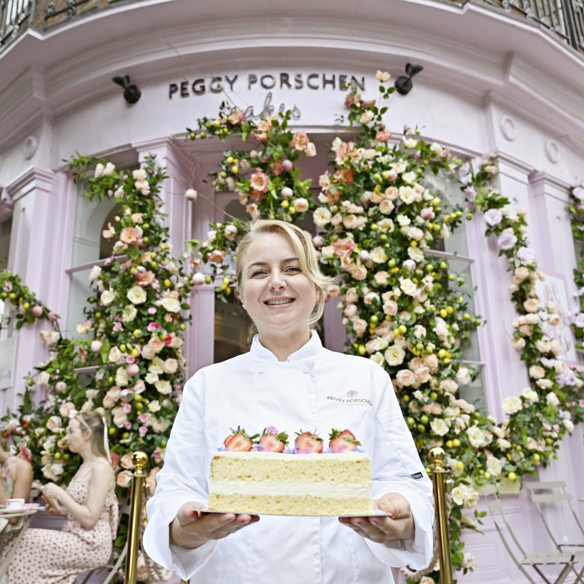 花で装飾された店舗前でケーキを持つペギー・ポーション氏